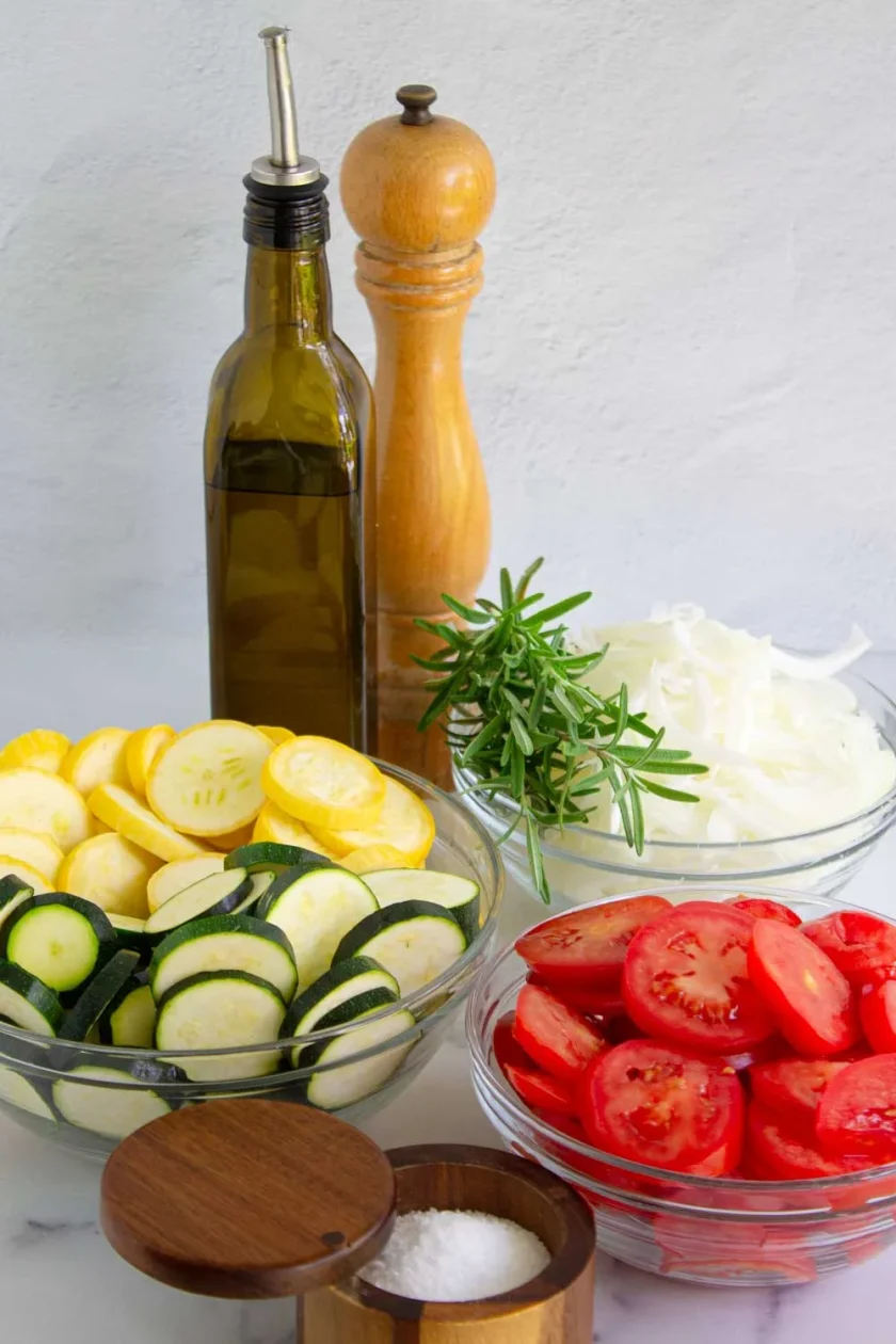 Bowls of sliced zucchini, yellow squash, tomatoes, and onions are arranged with olive oil, a pepper grinder, a small bowl of salt, and a sprig of rosemary on a white surface.