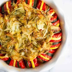 A round baking dish filled with sliced zucchini, yellow squash, and tomato, topped with cooked onions and herbs, arranged in a circular pattern.