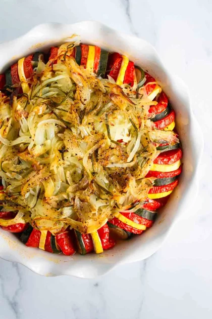 A round baking dish filled with sliced zucchini, yellow squash, and tomato, topped with cooked onions and herbs, arranged in a circular pattern.