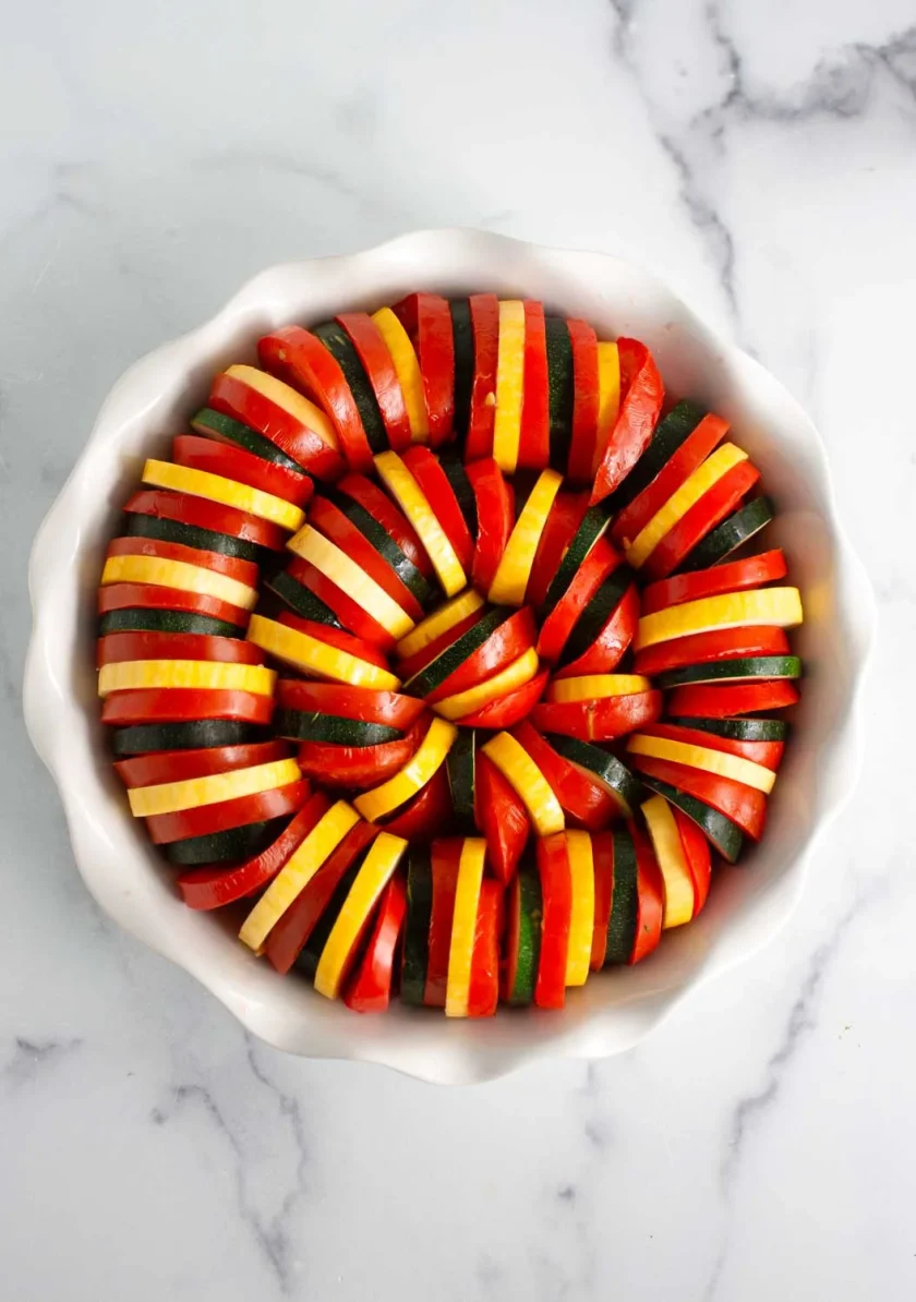 Rodajas de tomate, calabacín y calabaza amarilla dispuestas en círculos concéntricos en una fuente de horno blanca sobre una superficie de mármol.