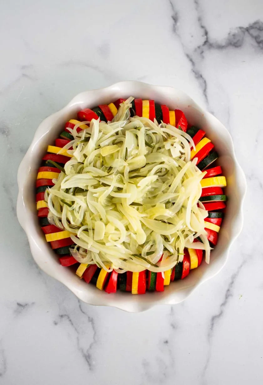 A round baking dish with layered slices of zucchini, yellow squash, and tomato arranged in a circle, topped with cooked onions, on a marble surface.