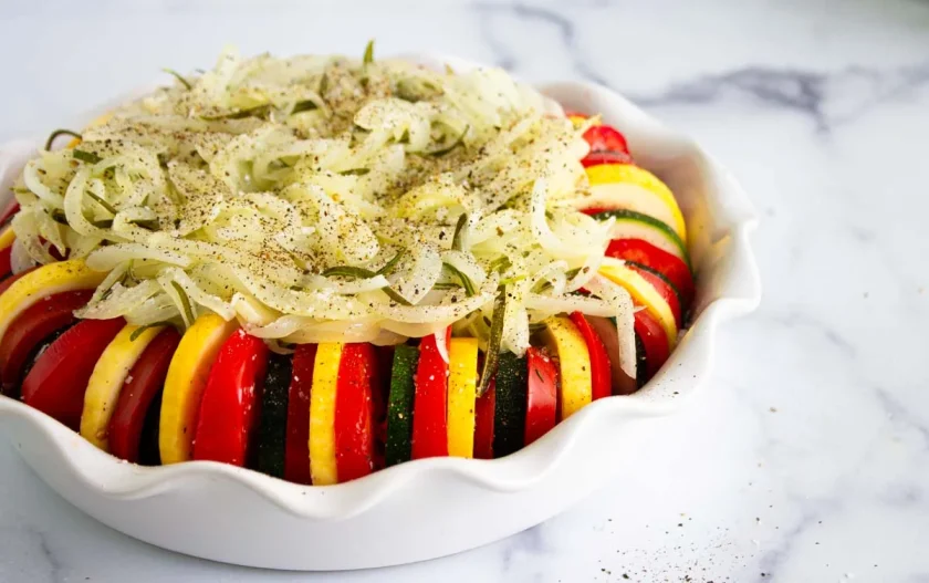 Sliced zucchini, yellow squash, and tomato arranged in a round baking dish, topped with seasoned onions and pepper, on a marble surface.