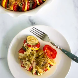 A serving of baked ratatouille with sliced tomatoes, zucchini, and onions is plated with a fork next to a baking dish on a white marble surface.