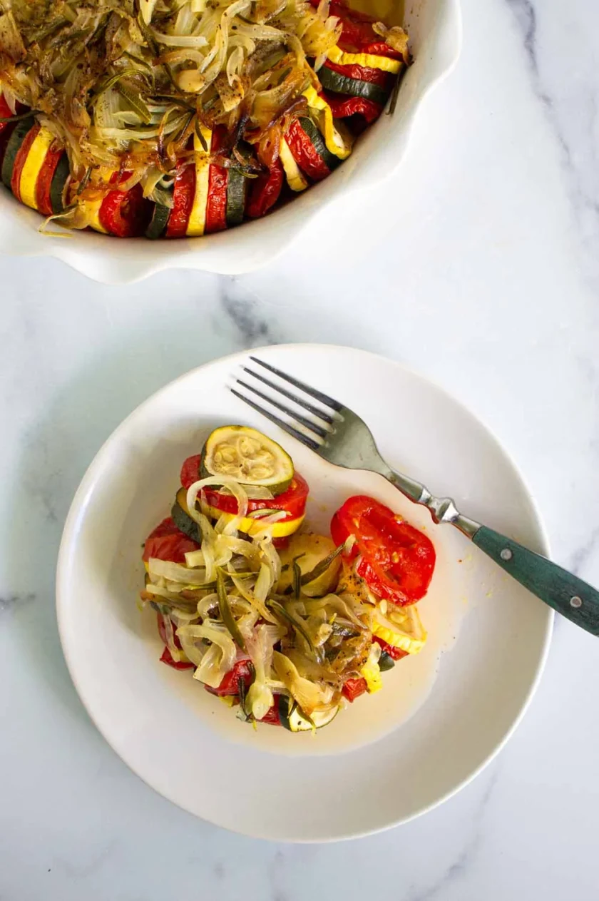 A serving of baked ratatouille with sliced tomatoes, zucchini, and onions is plated with a fork next to a baking dish on a white marble surface.