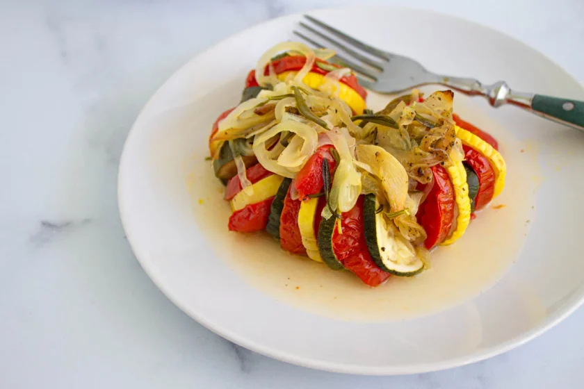 Un plato con una ración de rodajas de calabacín, calabaza amarilla, tomates y cebollas, dispuestas en capas, junto a un tenedor sobre una superficie blanca.