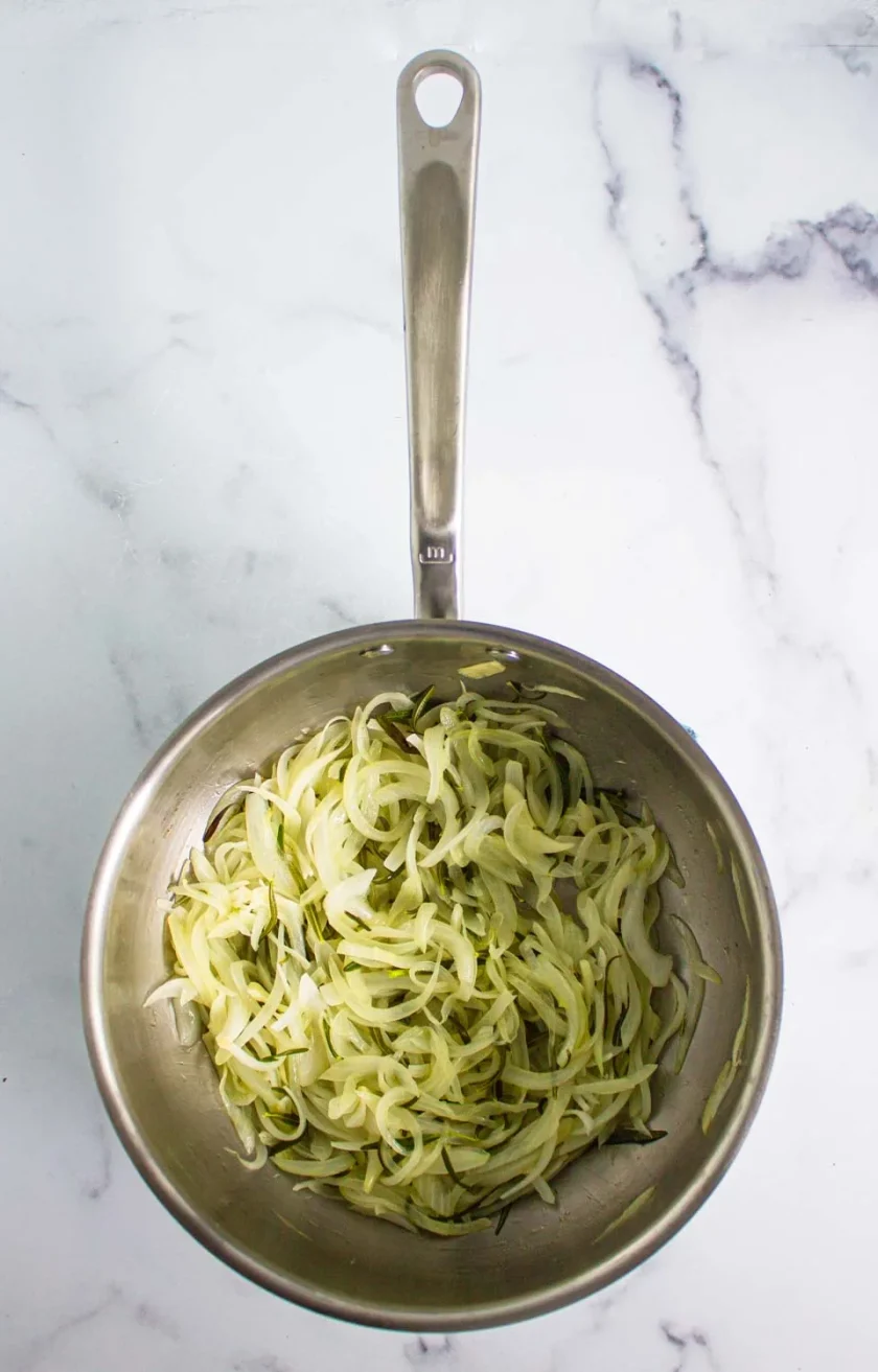 A stainless steel saucepan filled with cooked, thinly sliced onions sits on a white marble surface.