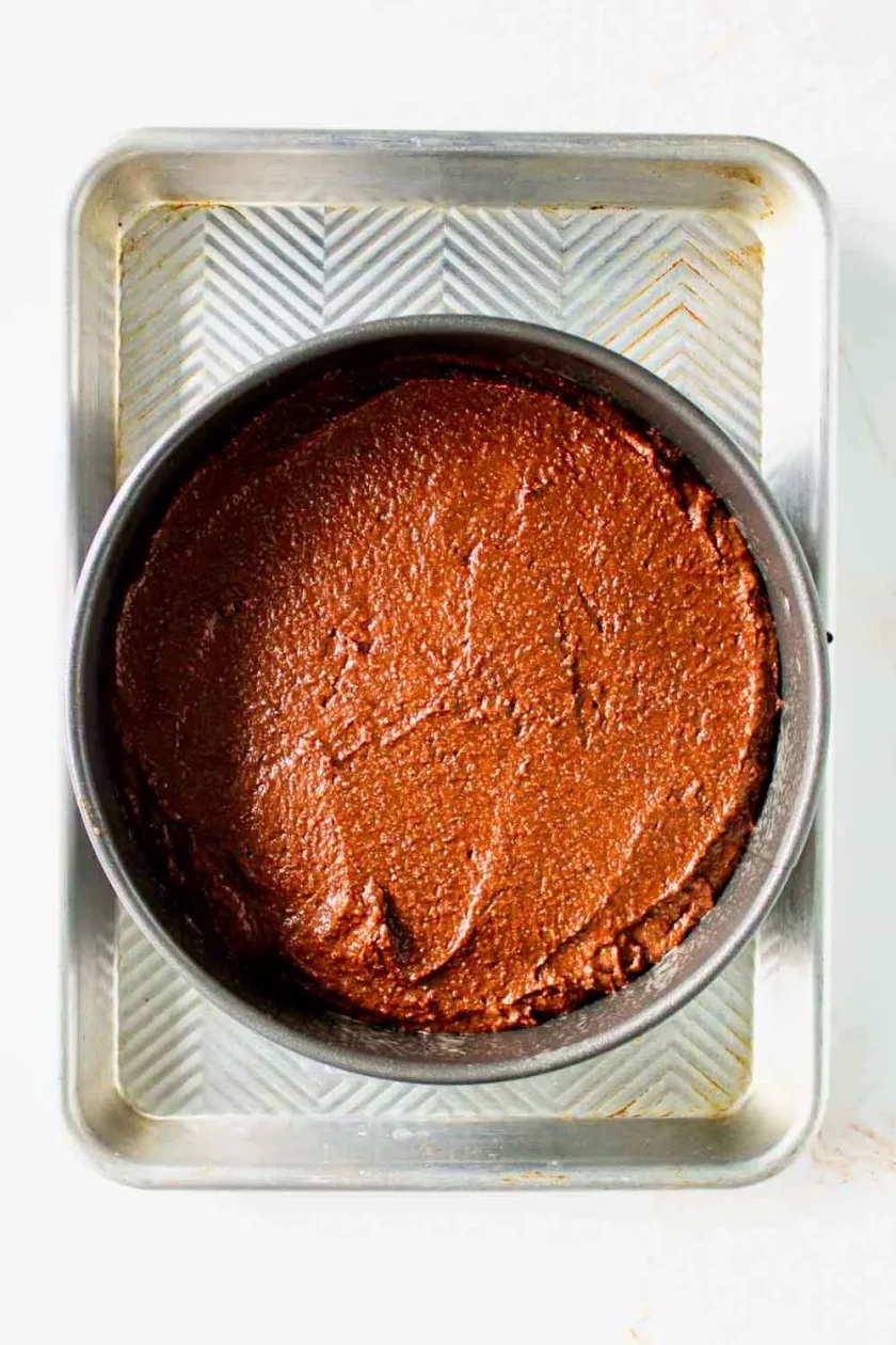 Un molde redondo lleno de masa para bizcocho de chocolate descansa dentro de una bandeja de horno metálica más grande, sobre una superficie blanca.
