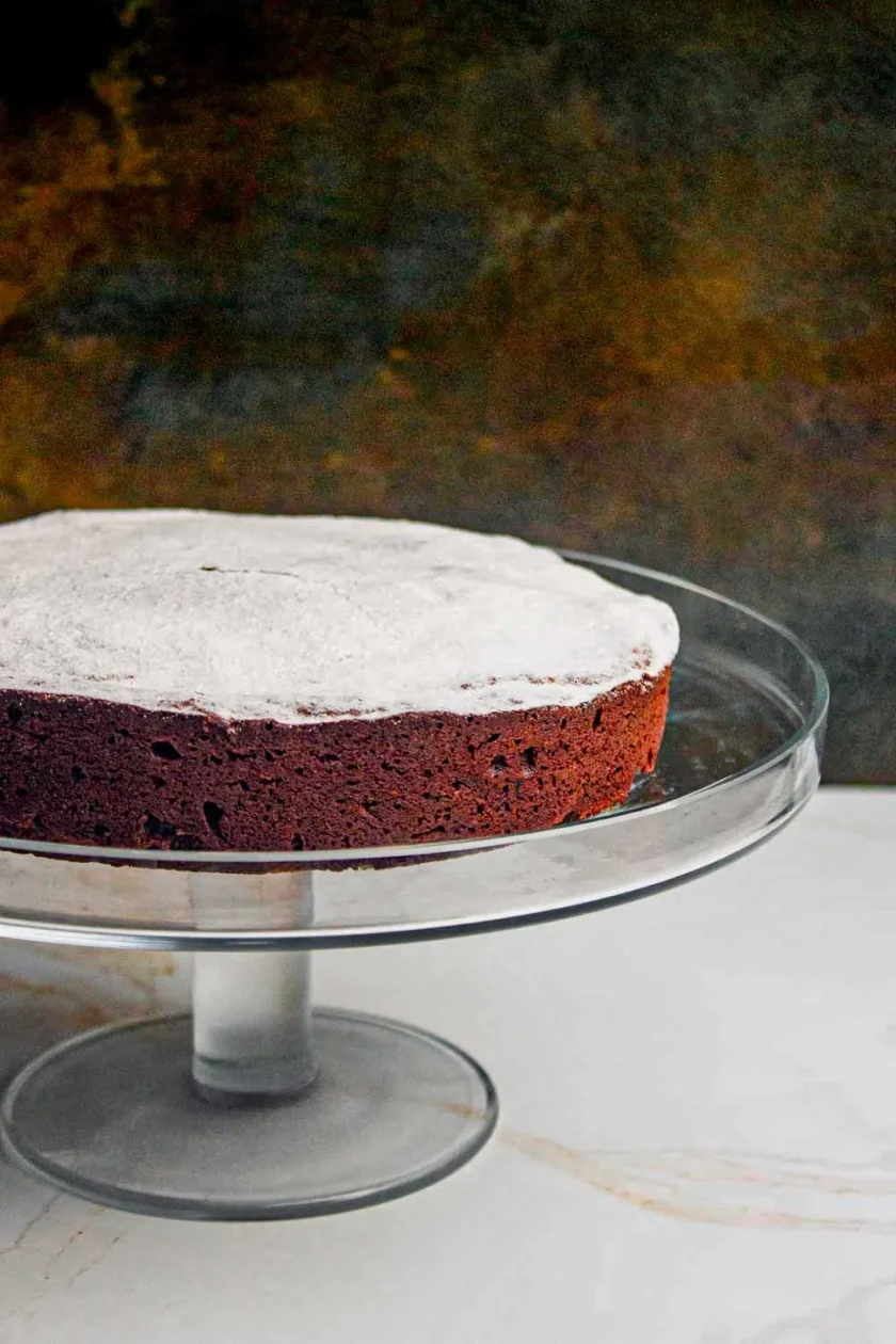 Una tarta redonda de chocolate, ligeramente espolvoreada con azúcar glas, descansa sobre un soporte de cristal para tartas sobre un fondo oscuro.
