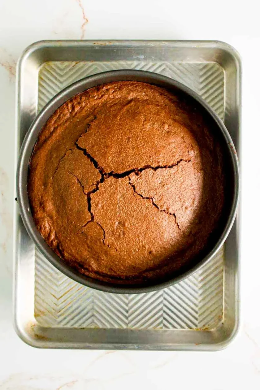 Un pastel redondo de chocolate con la superficie agrietada descansa en un molde metálico colocado sobre una bandeja de horno, fotografiado desde arriba.