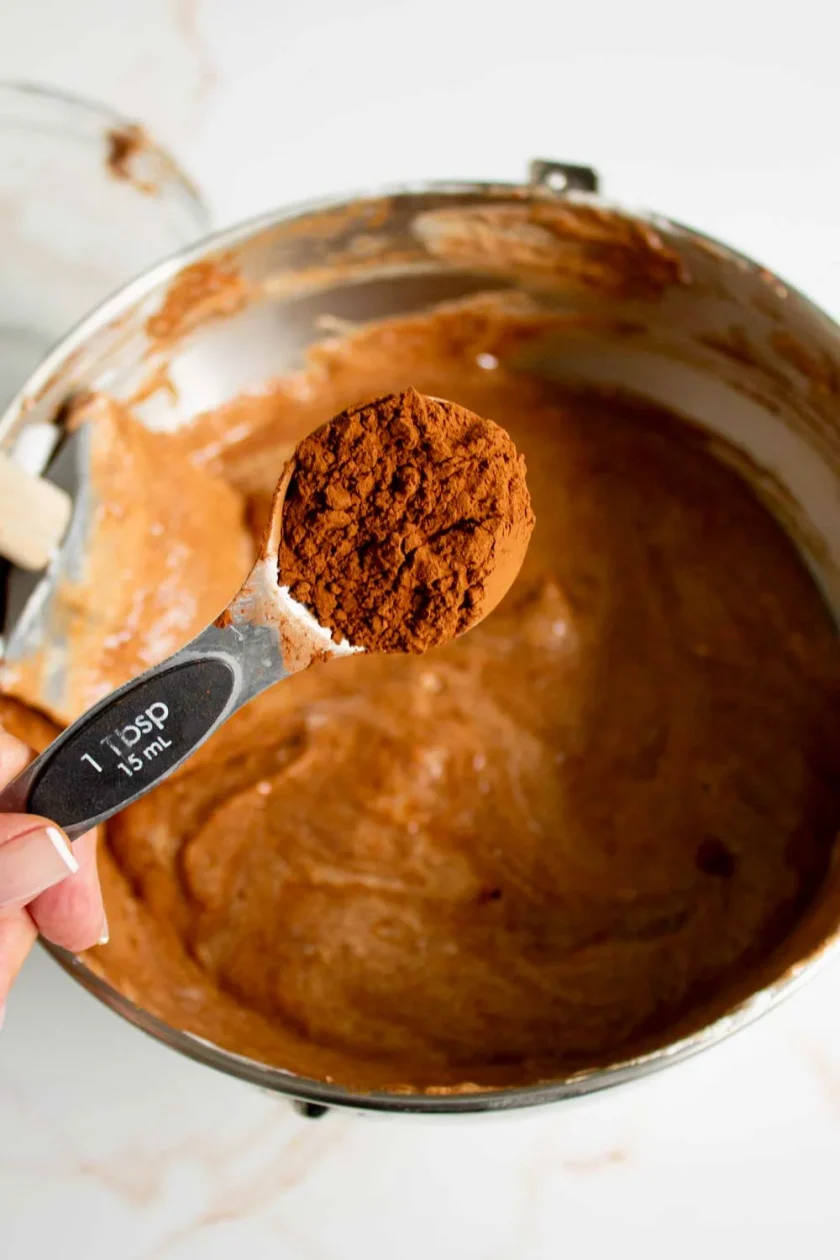 Una mano sostiene una cucharada de cacao en polvo sobre un bol lleno de masa de chocolate.