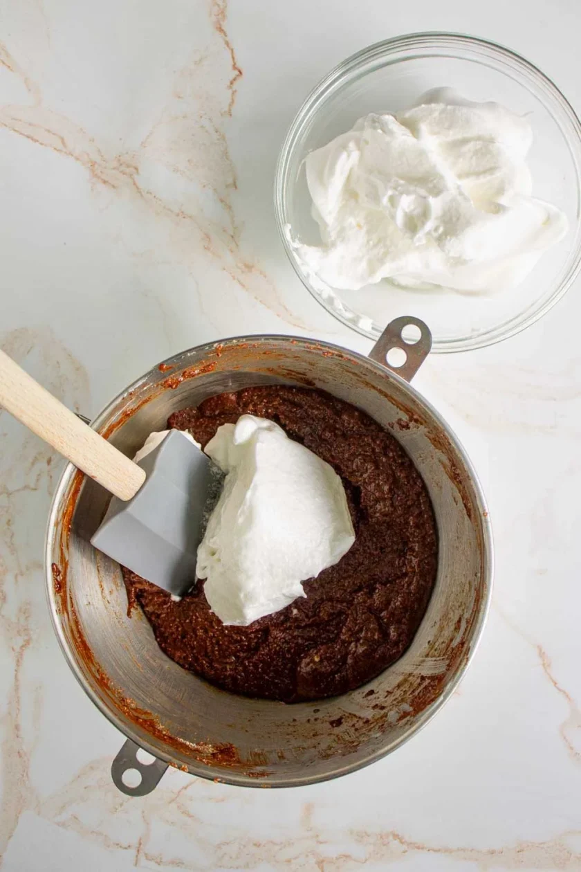 Un bol con masa de chocolate en el que se están incorporando claras de huevo batidas con una espátula, junto a otro bol con más claras de huevo batidas sobre una encimera de mármol.
