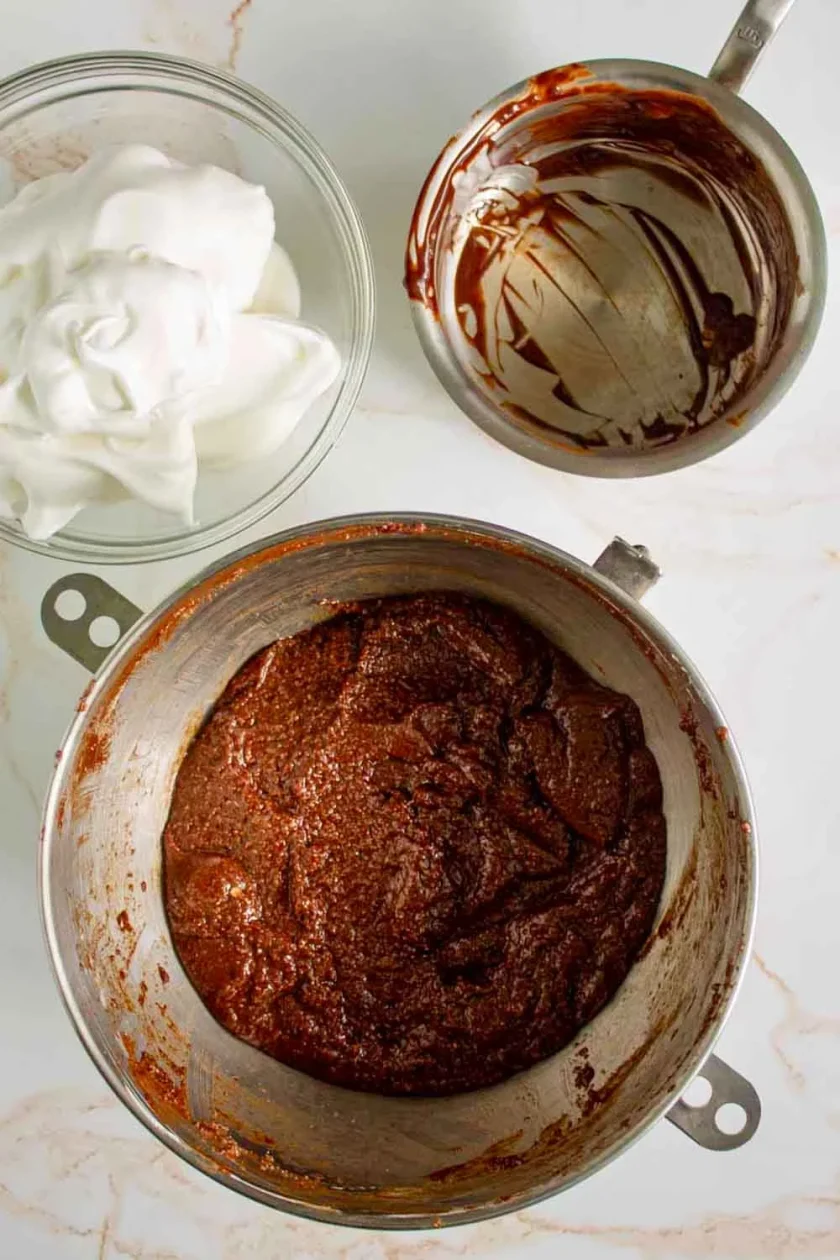 Sobre una encimera de mármol hay un bol con claras de huevo batidas, un bol vacío manchado de chocolate y un bol grande con masa de chocolate.