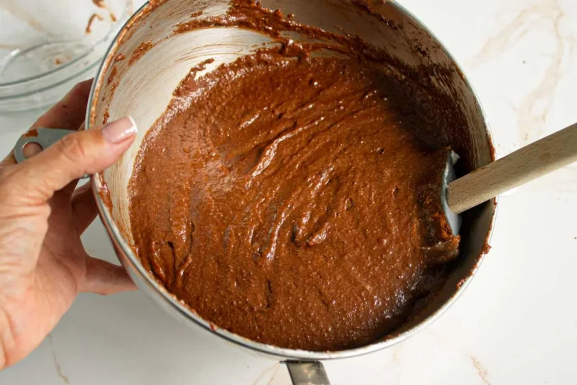 Una mano sostiene un bol metálico lleno de masa de chocolate, con una espátula parcialmente sumergida en la mezcla.
