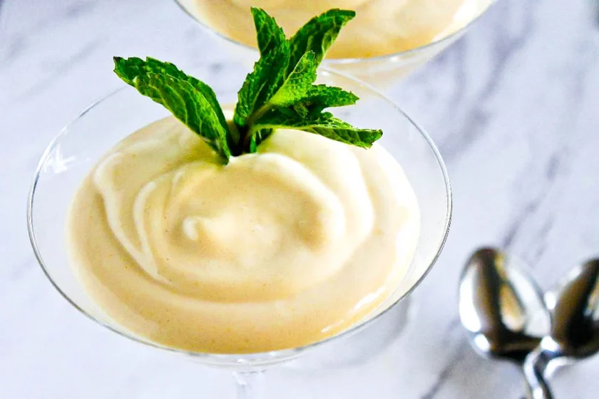 Un plato de cristal lleno de un postre cremoso, adornado con hojas de menta fresca, está colocado sobre una superficie de mármol junto a dos cucharas de metal.