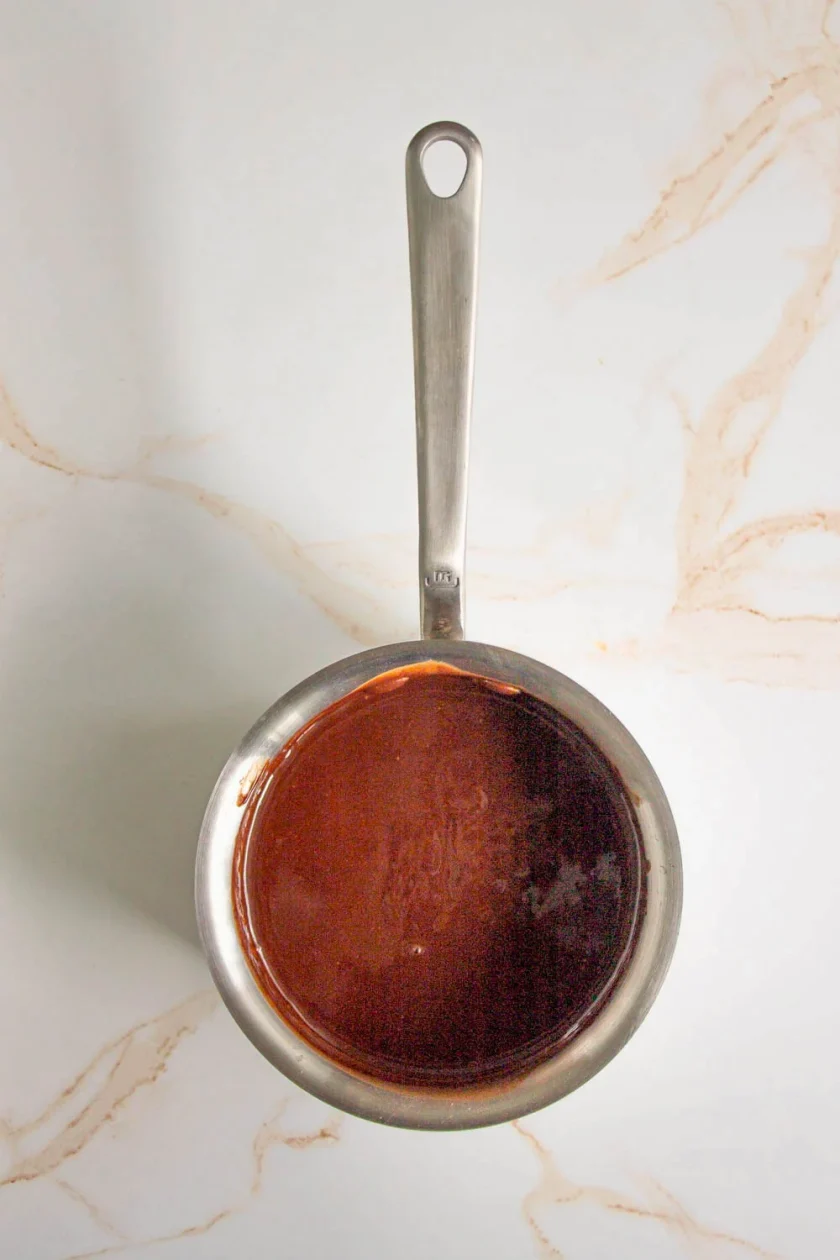 Una cacerola de acero inoxidable llena de salsa de chocolate está colocada sobre una superficie de mármol blanco con vetas de color marrón claro.