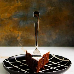 A slice of chocolate cake from una receta súper fácil de torta caprese, with a fork inserted, served on a black plate with a white crisscross pattern against a textured background.