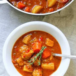 Un cuenco blanco con un sustancioso estofado de cerdo con zanahorias, papas y una ramita de tomillo, colocado sobre un mantel a rayas junto a una olla con el mismo guiso.