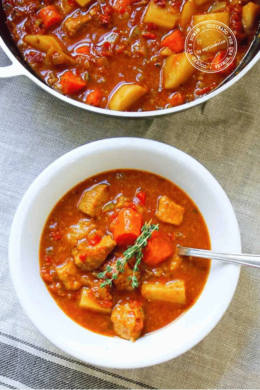 Un cuenco blanco con un sustancioso estofado de cerdo con zanahorias, papas y una ramita de tomillo, colocado sobre un mantel a rayas junto a una olla con el mismo guiso.