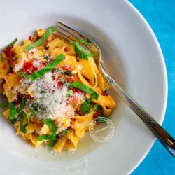 Un plato blanco de pasta, aderezado con queso rallado, albahaca fresca y pimientos rojos, descansa sobre una mesa azul con un tenedor al lado.