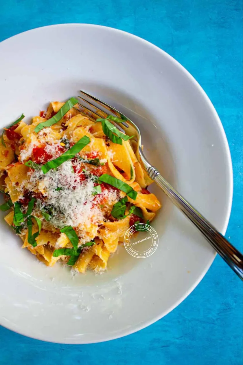 Un plato blanco de pasta, aderezado con queso rallado, albahaca fresca y pimientos rojos, descansa sobre una mesa azul con un tenedor al lado.