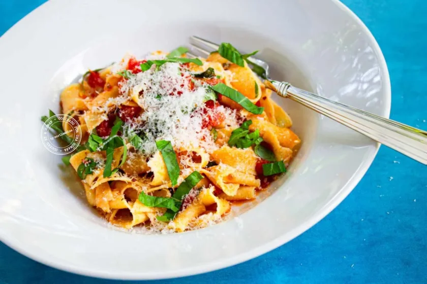 Un cuenco blanco lleno de pasta con salsa de tomate, queso rallado y albahaca fresca, colocado sobre una superficie azul con un tenedor apoyado en el borde del cuenco.