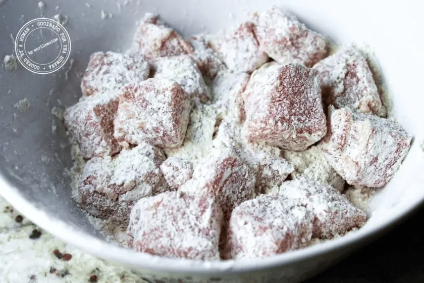 Se colocan cubos de lomo de cerdo cruda rebozados en maicena en un cuenco blanco, probablemente para prepararlos para un proceso de cocción, como dorarlos o guisarlos.