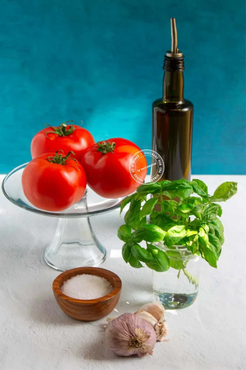Tres tomates sobre un soporte de cristal, una botella de aceite de oliva, un vaso con albahaca fresca, un cuenco con sal y una cabeza de ajo sobre una superficie blanca con fondo azul.