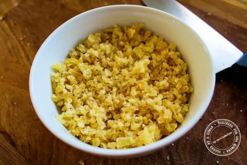 A white bowl filled with finely chopped crystallized ginger rests on a wooden surface next to a kitchen knife.