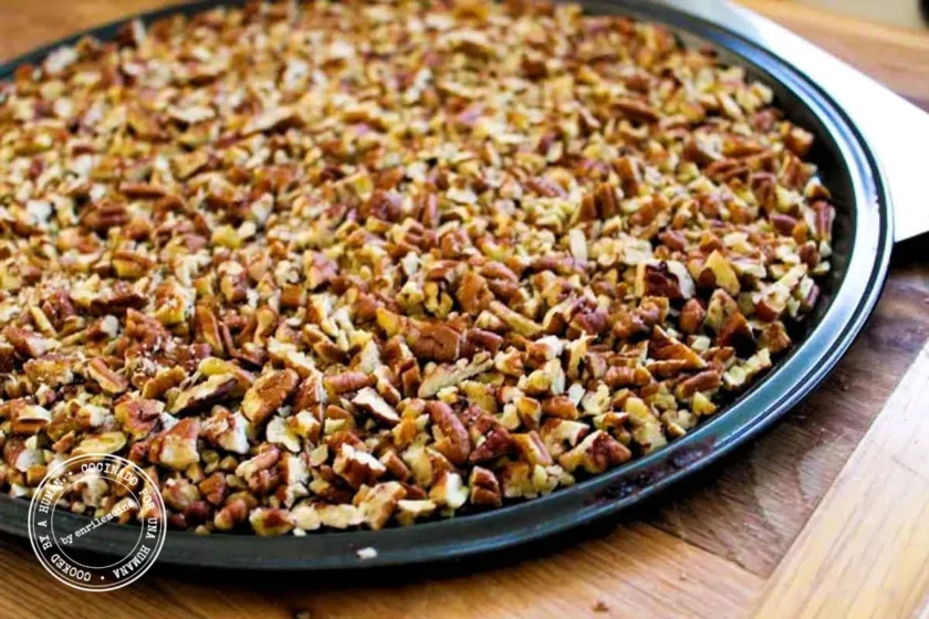 A round baking pan covered with an even layer of chopped pecans rests on a wooden surface.