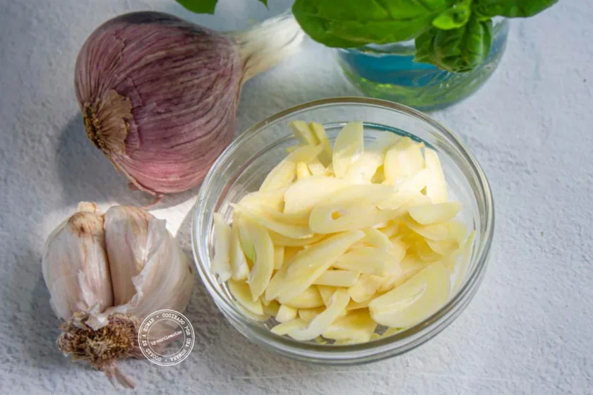 Un cuenco con ajos cortados en rodajas descansa junto a unas cabezas de ajo enteras y un frasco con albahaca fresca sobre una superficie clara.