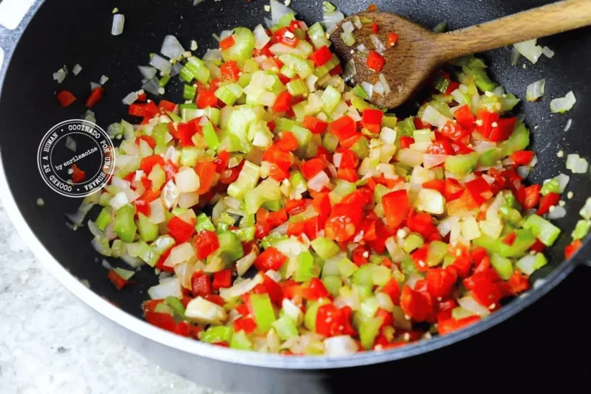 En la encimera de la cocina se están salteando pimientos rojos, apio y cebollas cortados en dados en caldero con una cuchara de madera.