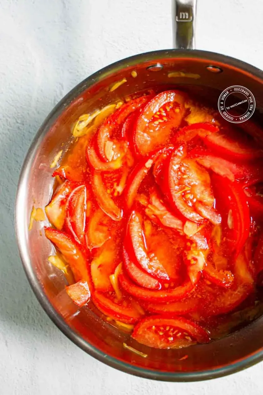 Los tomates y las cebollas cortados en rodajas se cocinan a fuego lento en una cacerola de acero inoxidable llena de líquido, sobre una superficie de color claro.