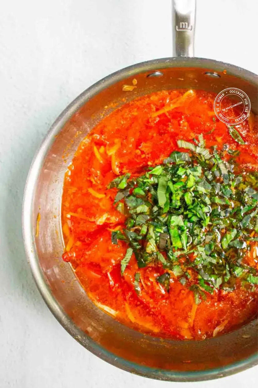 Una sartén de acero inoxidable llena de salsa de tomate roja, adornada con albahaca fresca picada, descansa sobre una superficie clara.