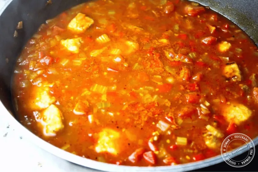 Un caldeto llena de un guiso a base de tomate con verduras cortadas en dados y trozos de cerdo, que se cocina a fuego lento en un caldo espeso y de color rojo.