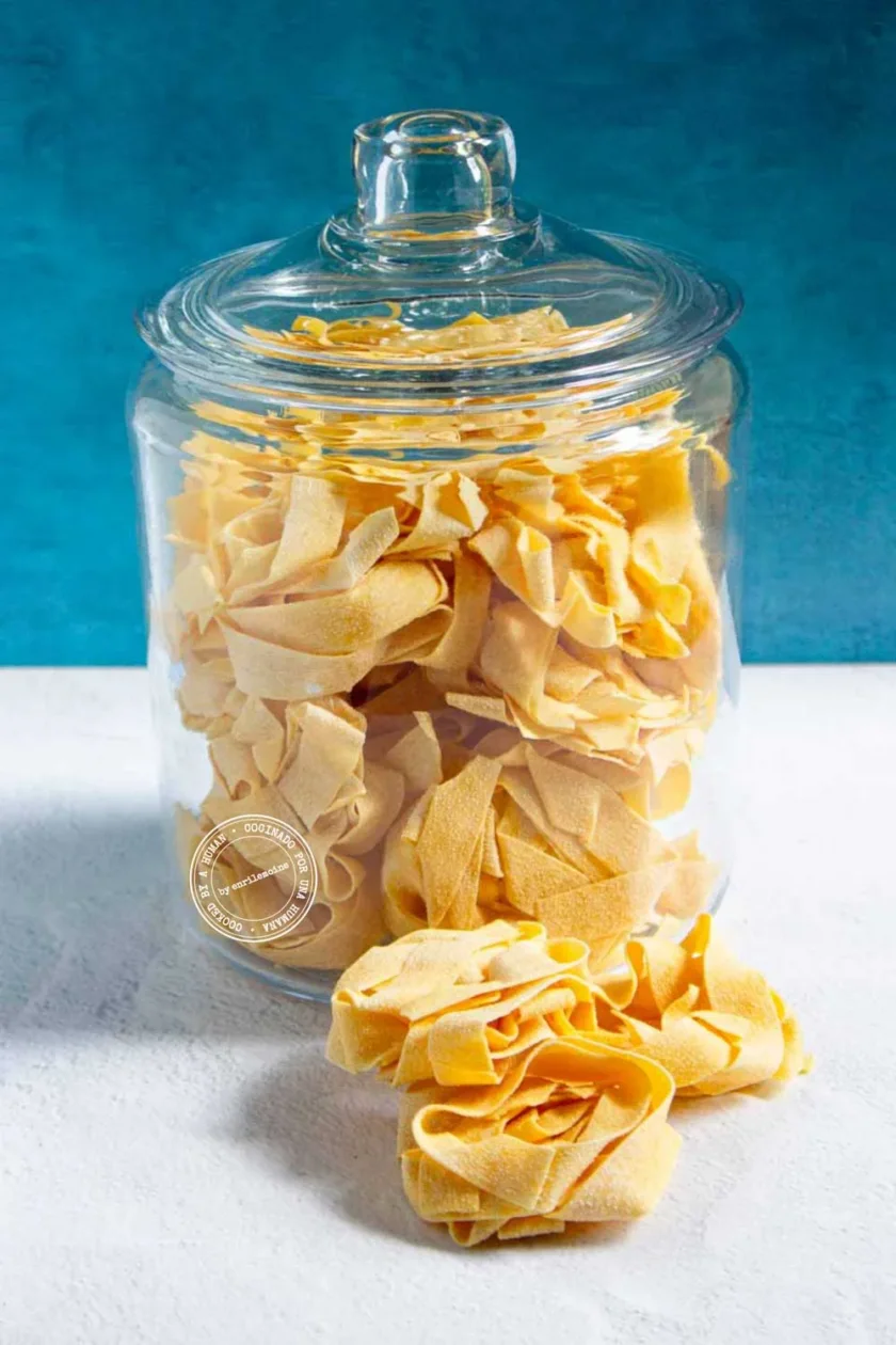 Un tarro de cristal lleno de pasta pappardelle cruda descansa sobre una superficie blanca, con varios nidos de pasta colocados fuera del tarro.