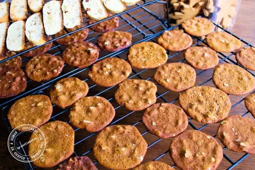 Rejillas de enfriamiento con filas de galletas planas y redondas con frutos secos y rodajas de biscotti encima, todas colocadas sobre una superficie de madera.