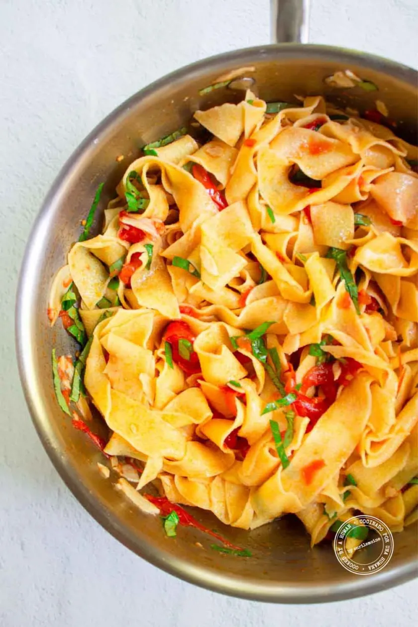 Anchas tiras de pasta salteadas con tomates y hierbas frescas en una sartén de acero inoxidable, vistas desde arriba.