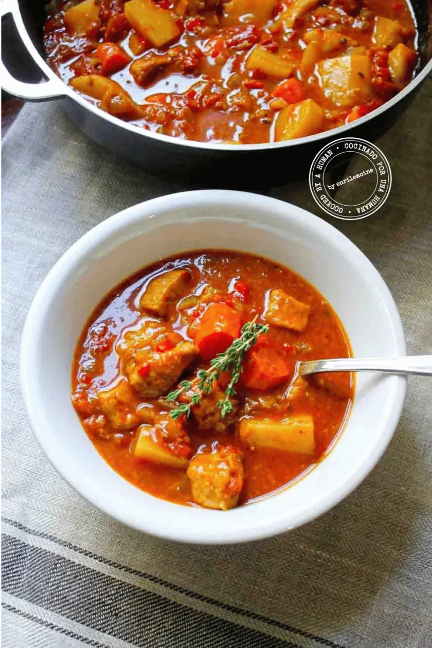 Un plato de un sustancioso estofado de cerdo tcon zanahorias, papas y una ramita de tomillo, con una cuchara en el plato y una olla de guiso al fondo.