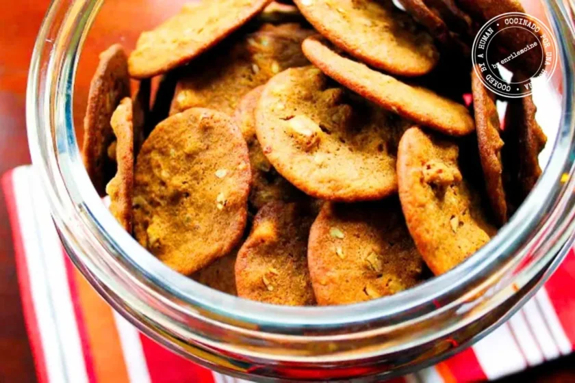 Un tarro de cristal lleno de galletas de jengibre en las que se aprecian trocitos de frutos secos, colocado sobre un paño a rayas que yace sobre una superficie de madera.