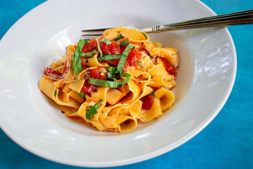 Un cuenco blanco con fideos anchos, coronado con tomates cortados en dados y albahaca fresca, con un tenedor apoyado en el borde, sobre un fondo azul.