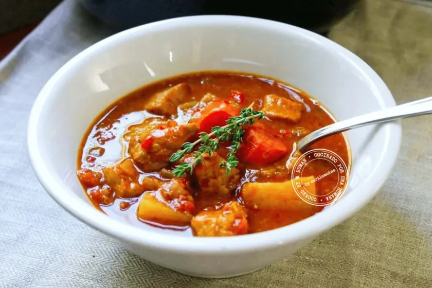 Un cuenco blanco lleno de estofado de cerdo con papas, zanahorias y hierbas, con una cuchara dentro.