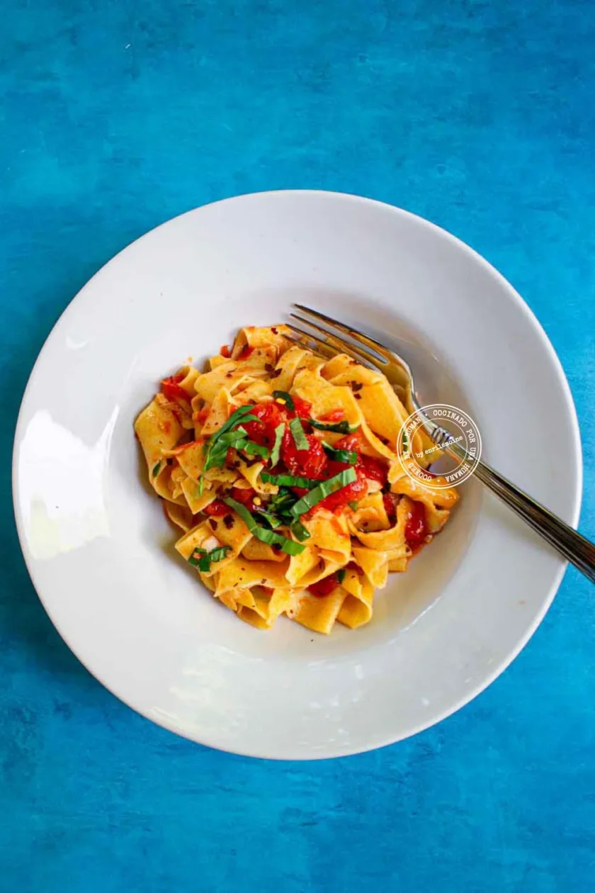 Un plato blanco con pasta ancha, cubierta con tomates cortados en dados y albahaca fresca, con un tenedor al lado, colocado sobre una superficie de color azul brillante.