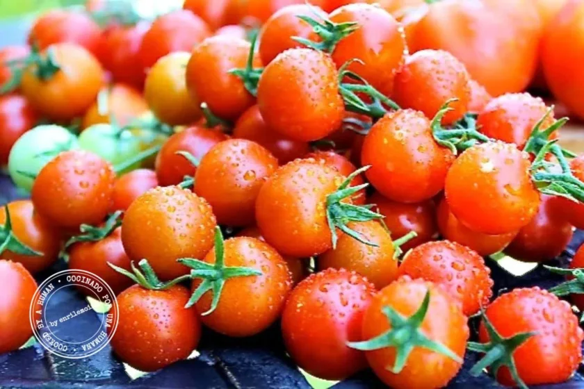 Un montón de tomates cherry frescos, con gotitas de agua en la piel, dispuestos sobre una superficie oscura, pone de manifiesto su frescura recién cosechada: un consejo sencillo para «alargarle la vida a los tomates» es mantenerlos secos y frescos. En la esquina inferior izquierda se ve un sello de certificación azul y blanco.