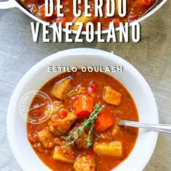 Un cuenco de estofado venezolano de cerdo con verduras y una ramita de tomillo, junto a una olla con el mismo estofado, sobre un mantel a rayas.