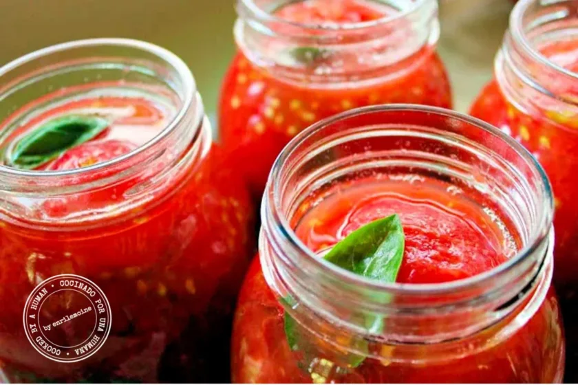 Cuatro tarros de cristal llenos de tomates pelados enteros en jugo de tomate, cada uno coronado con una hoja de albahaca fresca, listos para conservar.