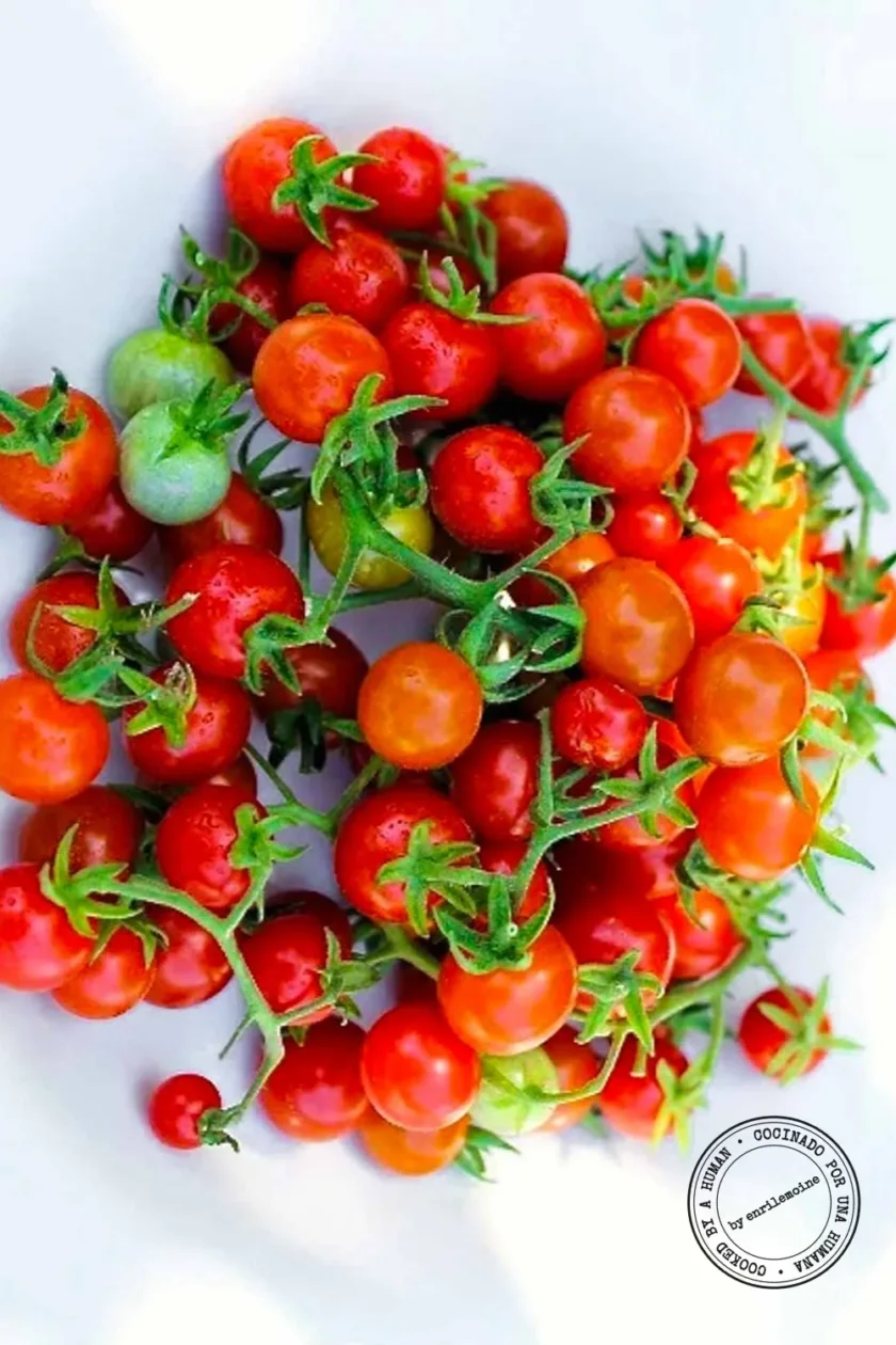Un montón de tomates cherry pequeños rojos y unos cuantos verdes, con el tallo aún intacto, expuestos sobre un fondo blanco.