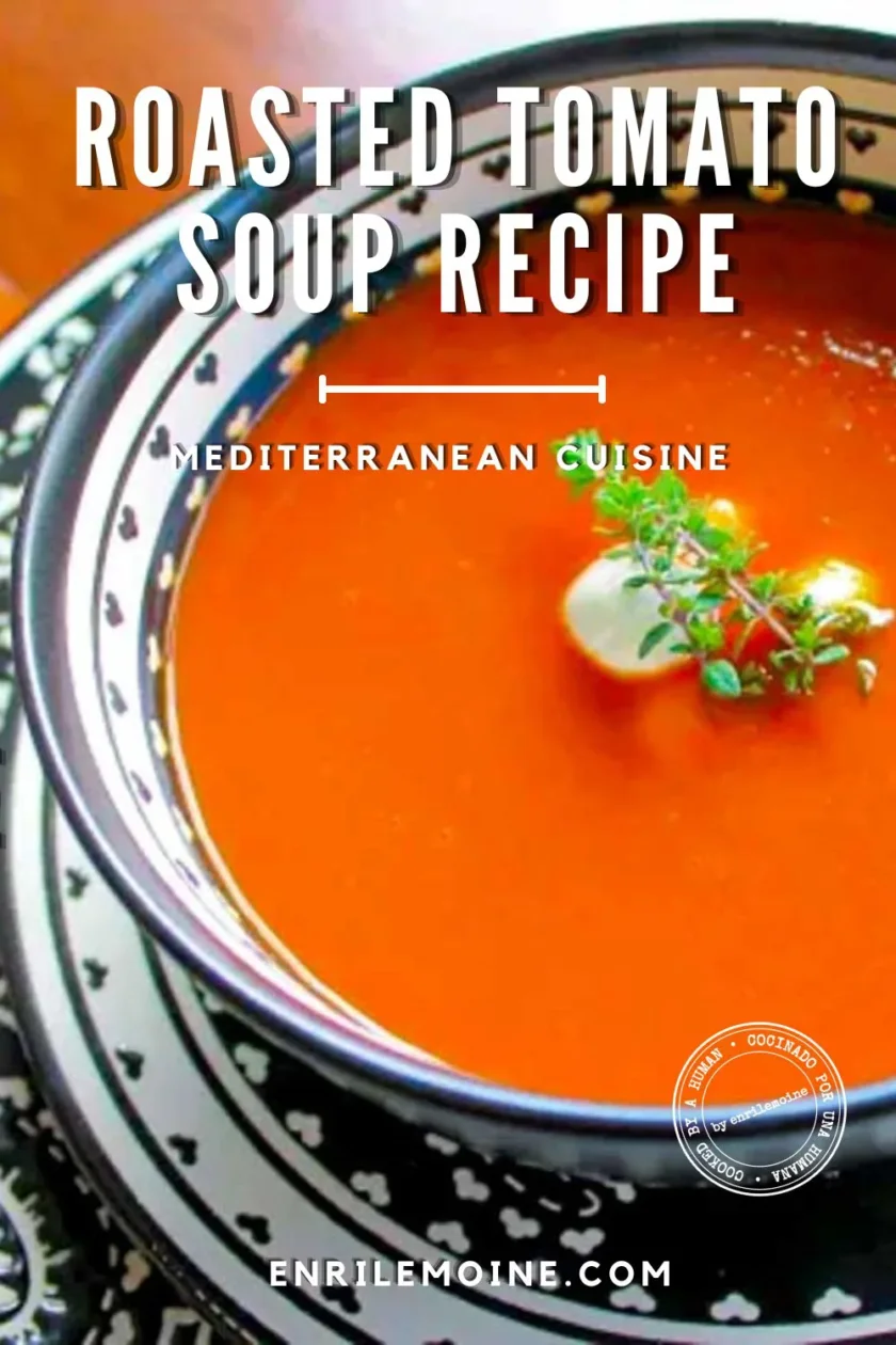 A bowl of roasted tomato soup with little balls of fresh goat cheese, garnished with fresh herbs, served on a plate with black-and-white arabesques, with a spoon on the side.