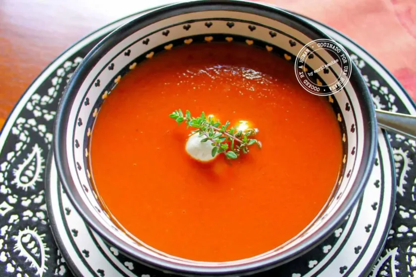 A bowl of roasted tomato soup with little balls of fresh goat cheese, garnished with fresh thyme, served on a plate with black-and-white arabesques, with a spoon on the side.