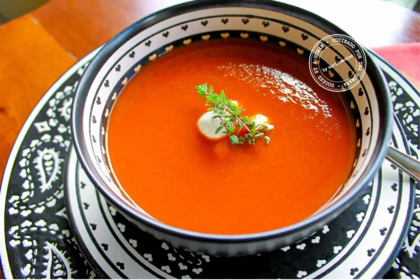 A bowl of roasted tomato soup with little balls of fresh goat cheese, garnished with fresh thyme, served on a plate with black-and-white arabesques, with a spoon on the side.