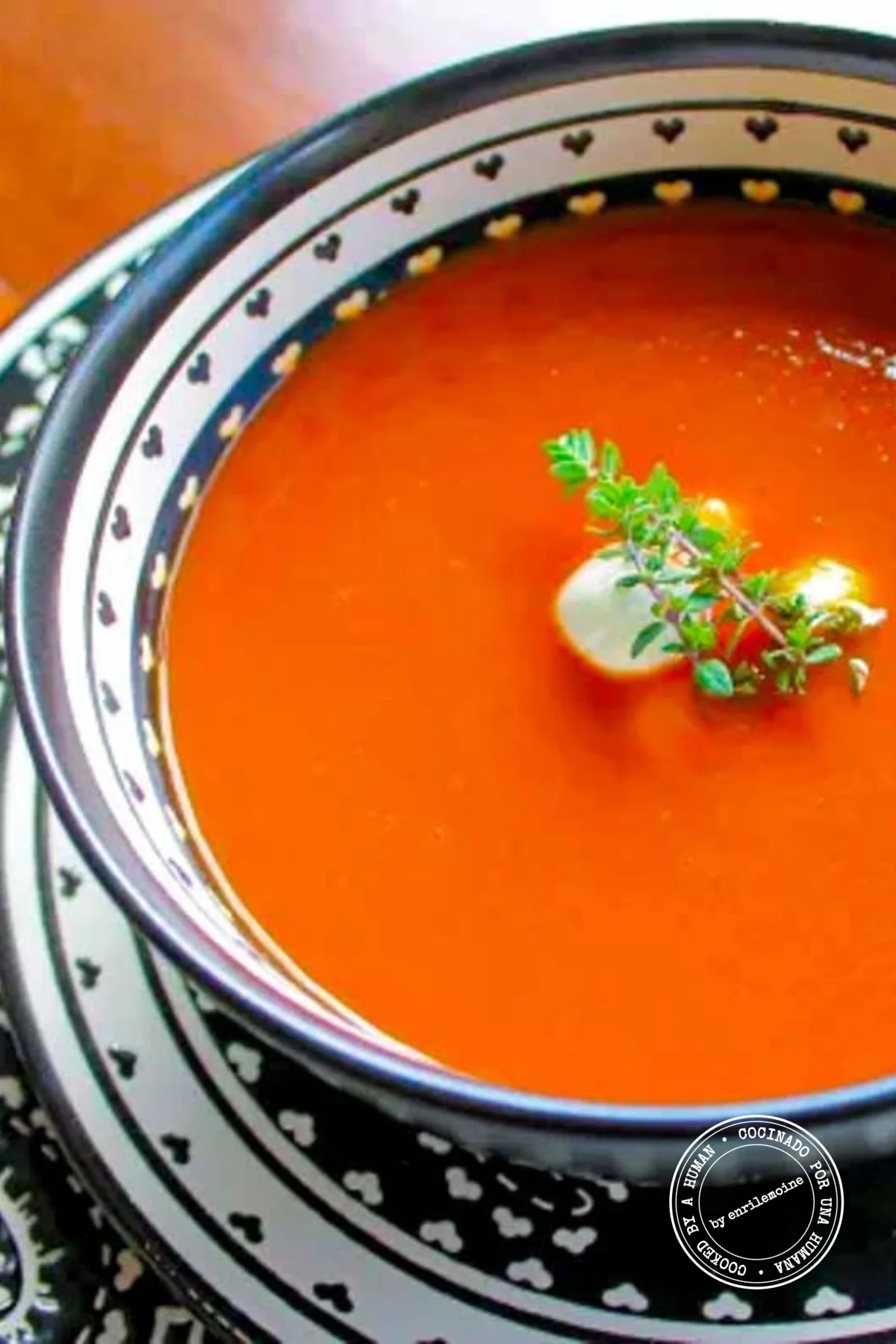 Un plato de sopa de tomate adornado con unas bolitas de queso de cabra fresco, una ramita de tomillo y un trocito de tomate, servido en un cuenco decorativo en blanco y negro.